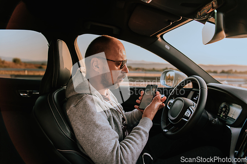 Image of A man with a sunglasses driving a car and type a message on smartphone at sunset. The concept of car travel