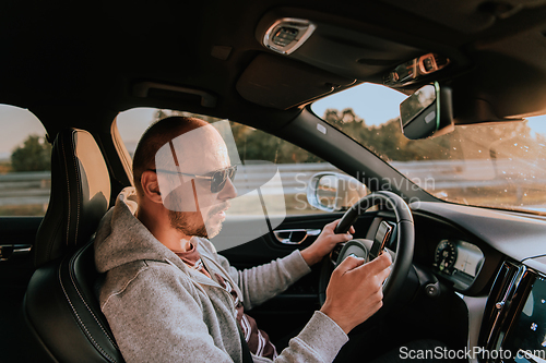 Image of A man with a sunglasses driving a car and type a message on smartphone at sunset. The concept of car travel