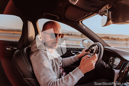 Image of A man with a sunglasses driving a car and type a message on smartphone at sunset. The concept of car travel