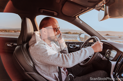 Image of A man with a sunglasses driving a car and talking on smartphone at sunset. The concept of car travel