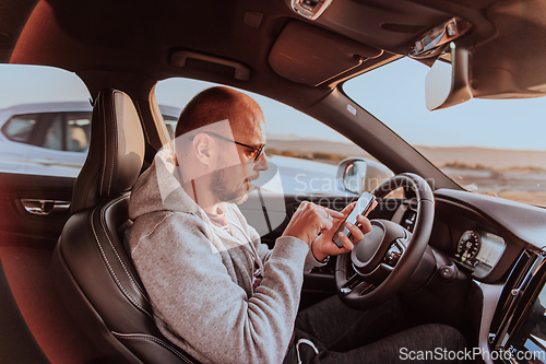 Image of A man with a sunglasses driving a car and type a message on smartphone at sunset. The concept of car travel