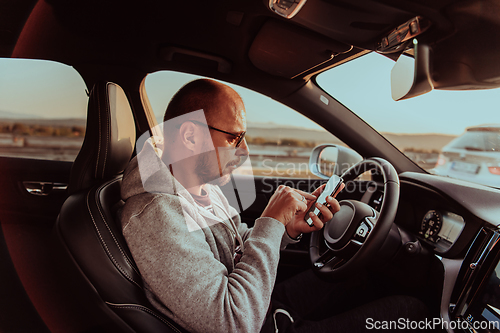 Image of A man with a sunglasses driving a car and type a message on smartphone at sunset. The concept of car travel