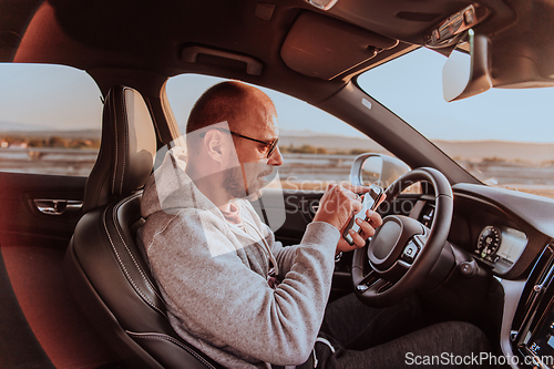 Image of A man with a sunglasses driving a car and type a message on smartphone at sunset. The concept of car travel