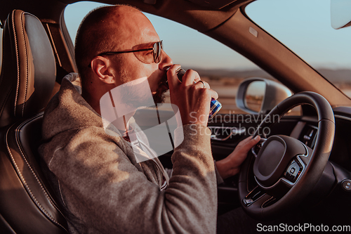 Image of A tired man drinking acoffee while driving a car at sunset. Tired travel and long drive