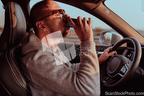Image of A tired man drinking acoffee while driving a car at sunset. Tired travel and long drive