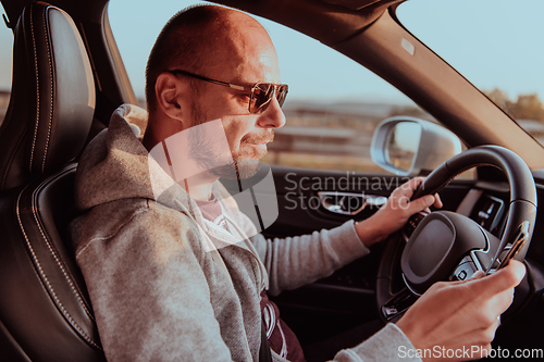 Image of A man with a sunglasses driving a car and type a message on smartphone at sunset. The concept of car travel