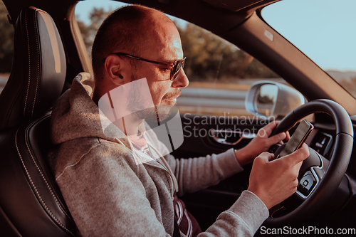 Image of A man with a sunglasses driving a car and type a message on smartphone at sunset. The concept of car travel