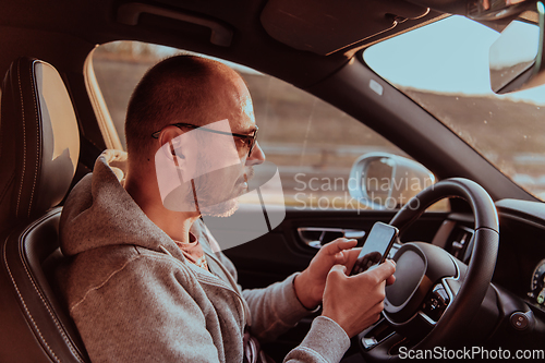 Image of A man with a sunglasses driving a car and type a message on smartphone at sunset. The concept of car travel