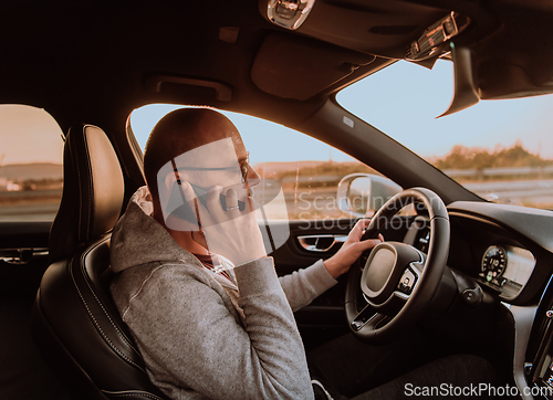 Image of A man with a sunglasses driving a car and talking on smartphone at sunset. The concept of car travel