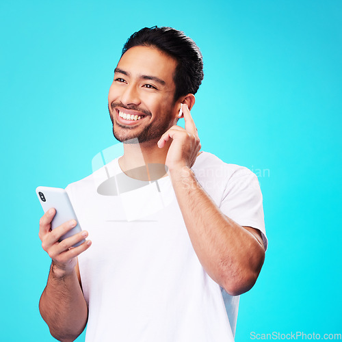 Image of Asian man, earphones and smartphone in studio for music, podcast app and blue background. Happy male model thinking with mobile phone, listening to audio and streaming sound of radio media with tech