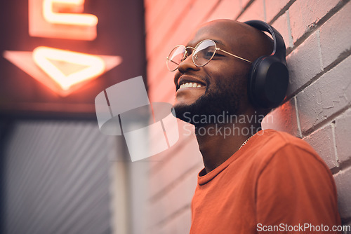 Image of .Thinking, smile and music with a black man in the city, leaning on a brick wall on the street at night. Idea, glasses and headphones with a happy young male person streaming or listening to audio.