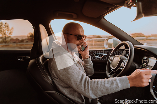 Image of A man with a sunglasses driving a car and talking on smartphone at sunset. The concept of car travel