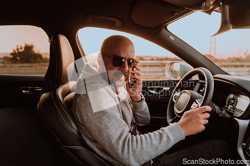 Image of A man with a sunglasses driving a car and talking on smartphone at sunset. The concept of car travel
