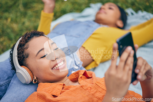 Image of Music, phone and lesbian couple relax at picnic on grass, technology and streaming service app in nature. Cellphone, listening to headphones and happy lgbt women on blanket in garden together in park