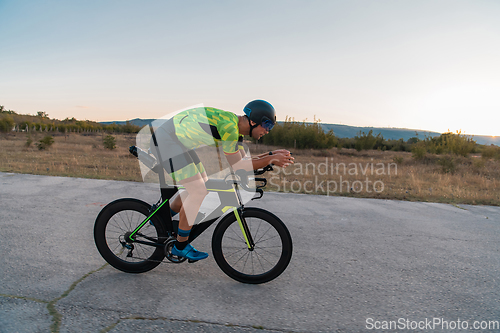 Image of Triathlete riding his bicycle during sunset, preparing for a marathon. The warm colors of the sky provide a beautiful backdrop for his determined and focused effort.