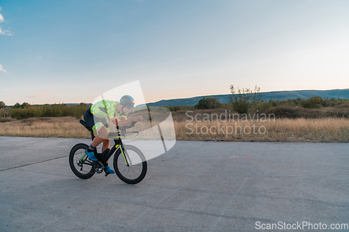 Image of Triathlete riding his bicycle during sunset, preparing for a marathon. The warm colors of the sky provide a beautiful backdrop for his determined and focused effort.