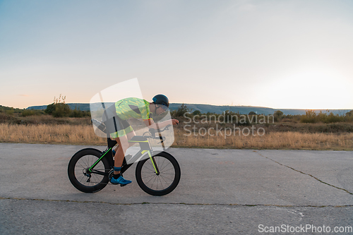 Image of Triathlete riding his bicycle during sunset, preparing for a marathon. The warm colors of the sky provide a beautiful backdrop for his determined and focused effort.