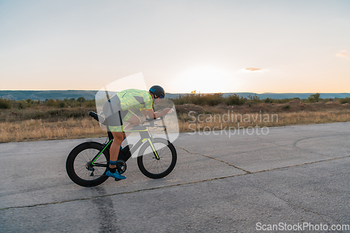 Image of Triathlete riding his bicycle during sunset, preparing for a marathon. The warm colors of the sky provide a beautiful backdrop for his determined and focused effort.