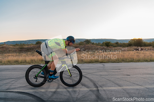 Image of Triathlete riding his bicycle during sunset, preparing for a marathon. The warm colors of the sky provide a beautiful backdrop for his determined and focused effort.