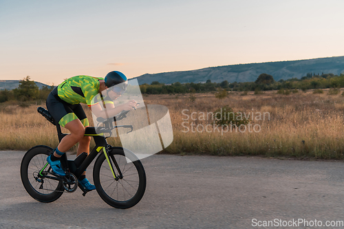 Image of Triathlete riding his bicycle during sunset, preparing for a marathon. The warm colors of the sky provide a beautiful backdrop for his determined and focused effort.