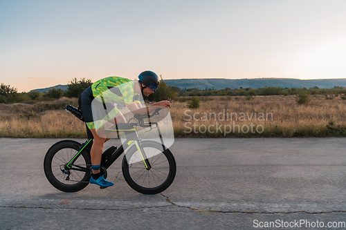 Image of Triathlete riding his bicycle during sunset, preparing for a marathon. The warm colors of the sky provide a beautiful backdrop for his determined and focused effort.