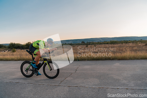 Image of Triathlete riding his bicycle during sunset, preparing for a marathon. The warm colors of the sky provide a beautiful backdrop for his determined and focused effort.