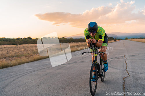 Image of Triathlete riding his bicycle during sunset, preparing for a marathon. The warm colors of the sky provide a beautiful backdrop for his determined and focused effort.