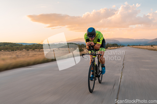 Image of Triathlete riding his bicycle during sunset, preparing for a marathon. The warm colors of the sky provide a beautiful backdrop for his determined and focused effort.