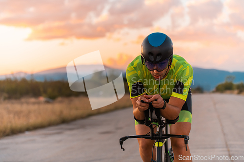 Image of Close up photo of triathlete riding his bicycle during sunset, preparing for a marathon. The warm colors of the sky provide a beautiful backdrop for his determined and focused effort.