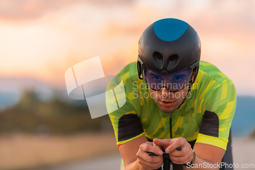 Image of Close up photo of triathlete riding his bicycle during sunset, preparing for a marathon. The warm colors of the sky provide a beautiful backdrop for his determined and focused effort.