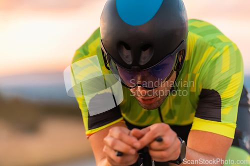 Image of Close up photo of triathlete riding his bicycle during sunset, preparing for a marathon. The warm colors of the sky provide a beautiful backdrop for his determined and focused effort.