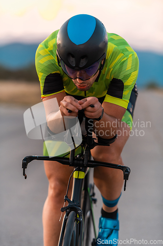 Image of Close up photo of triathlete riding his bicycle during sunset, preparing for a marathon. The warm colors of the sky provide a beautiful backdrop for his determined and focused effort.