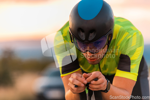 Image of Close up photo of triathlete riding his bicycle during sunset, preparing for a marathon. The warm colors of the sky provide a beautiful backdrop for his determined and focused effort.