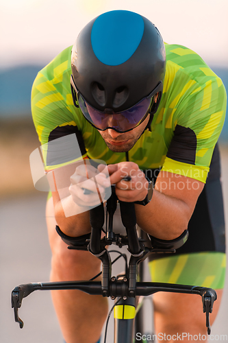 Image of Close up photo of triathlete riding his bicycle during sunset, preparing for a marathon. The warm colors of the sky provide a beautiful backdrop for his determined and focused effort.