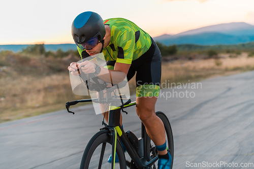 Image of Close up photo of triathlete riding his bicycle during sunset, preparing for a marathon. The warm colors of the sky provide a beautiful backdrop for his determined and focused effort.