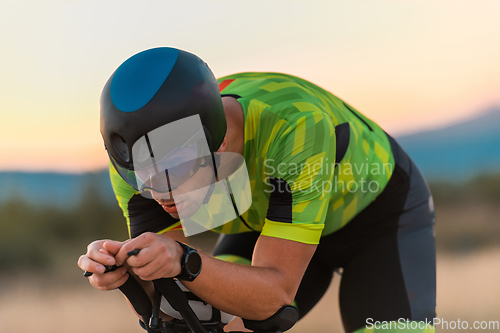 Image of Close up photo of triathlete riding his bicycle during sunset, preparing for a marathon. The warm colors of the sky provide a beautiful backdrop for his determined and focused effort.