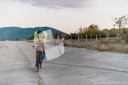 Image of Triathlete riding his bicycle during sunset, preparing for a marathon. The warm colors of the sky provide a beautiful backdrop for his determined and focused effort.