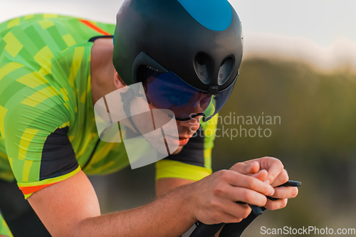 Image of Close up photo of triathlete riding his bicycle during sunset, preparing for a marathon. The warm colors of the sky provide a beautiful backdrop for his determined and focused effort.