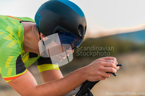 Image of Close up photo of triathlete riding his bicycle during sunset, preparing for a marathon. The warm colors of the sky provide a beautiful backdrop for his determined and focused effort.