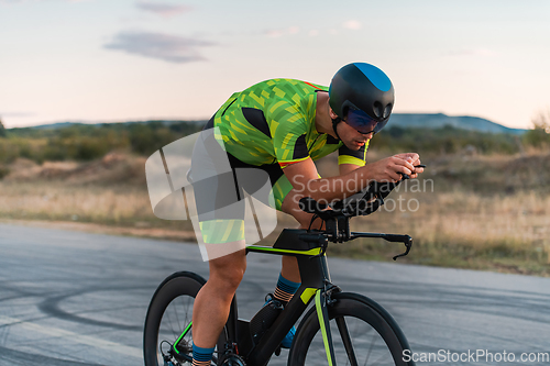 Image of Triathlete riding his bicycle during sunset, preparing for a marathon. The warm colors of the sky provide a beautiful backdrop for his determined and focused effort.