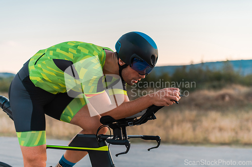 Image of Close up photo of triathlete riding his bicycle during sunset, preparing for a marathon. The warm colors of the sky provide a beautiful backdrop for his determined and focused effort.