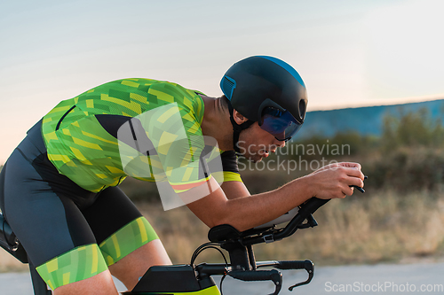 Image of Close up photo of triathlete riding his bicycle during sunset, preparing for a marathon. The warm colors of the sky provide a beautiful backdrop for his determined and focused effort.