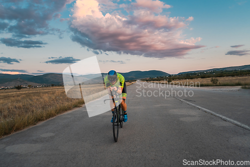 Image of Triathlete riding his bicycle during sunset, preparing for a marathon. The warm colors of the sky provide a beautiful backdrop for his determined and focused effort.