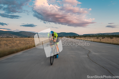 Image of Triathlete riding his bicycle during sunset, preparing for a marathon. The warm colors of the sky provide a beautiful backdrop for his determined and focused effort.