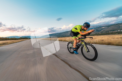 Image of Triathlete riding his bicycle during sunset, preparing for a marathon. The warm colors of the sky provide a beautiful backdrop for his determined and focused effort.