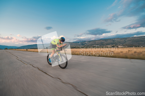 Image of Triathlete riding his bicycle during sunset, preparing for a marathon. The warm colors of the sky provide a beautiful backdrop for his determined and focused effort.