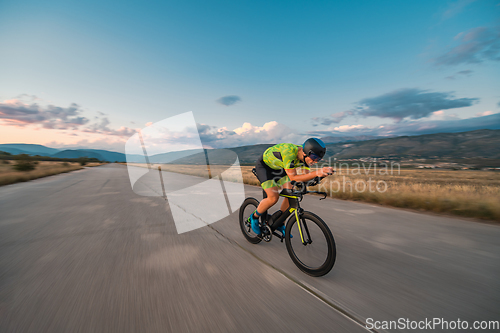 Image of Triathlete riding his bicycle during sunset, preparing for a marathon. The warm colors of the sky provide a beautiful backdrop for his determined and focused effort.