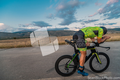 Image of Triathlete riding his bicycle during sunset, preparing for a marathon. The warm colors of the sky provide a beautiful backdrop for his determined and focused effort.