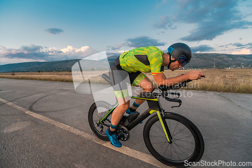 Image of Triathlete riding his bicycle during sunset, preparing for a marathon. The warm colors of the sky provide a beautiful backdrop for his determined and focused effort.