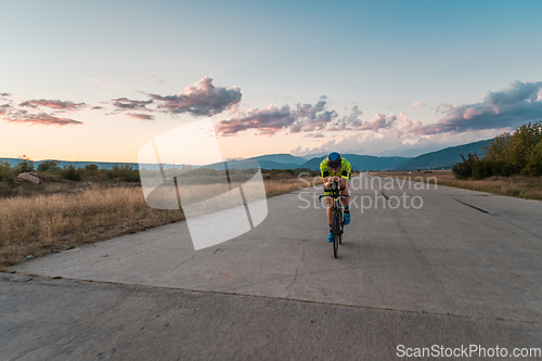 Image of Triathlete riding his bicycle during sunset, preparing for a marathon. The warm colors of the sky provide a beautiful backdrop for his determined and focused effort.